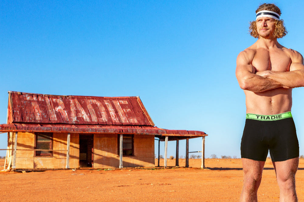 Nick Honey Badger Cummins athletic man in black trunks and headband standing in front of an old building in the outback.